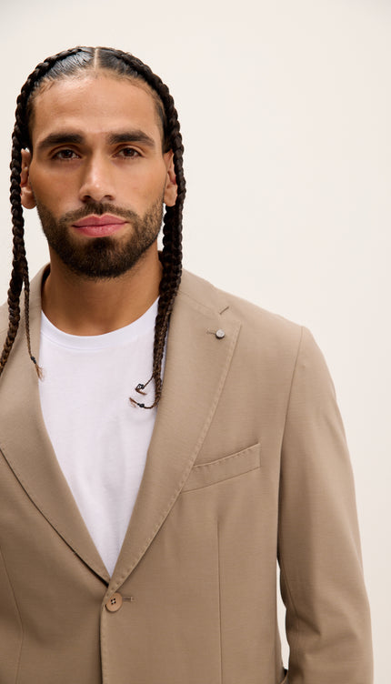 Man wearing a beige blazer over a white shirt with braided hair against a plain background