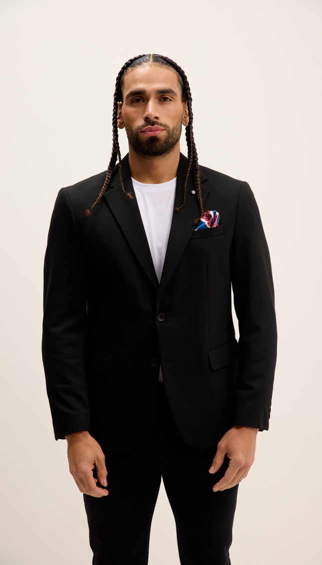 Man wearing a black suit with a white shirt and colorful lapel pin on a plain background