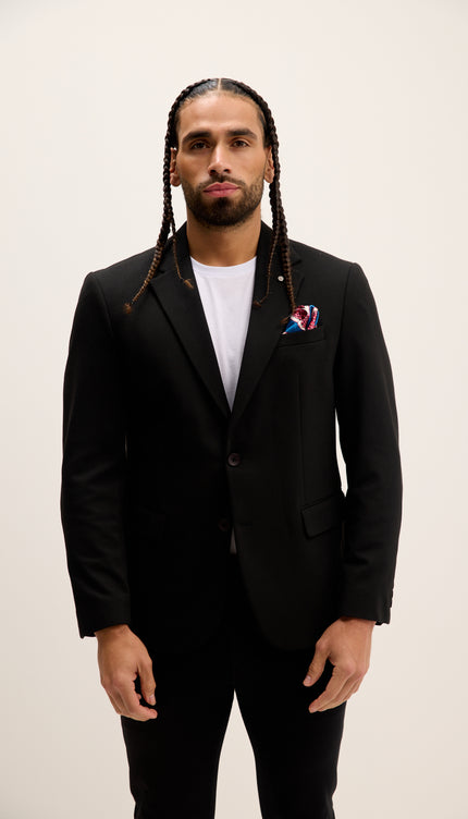 Man wearing a black suit with a white shirt and colorful lapel pin on a plain background